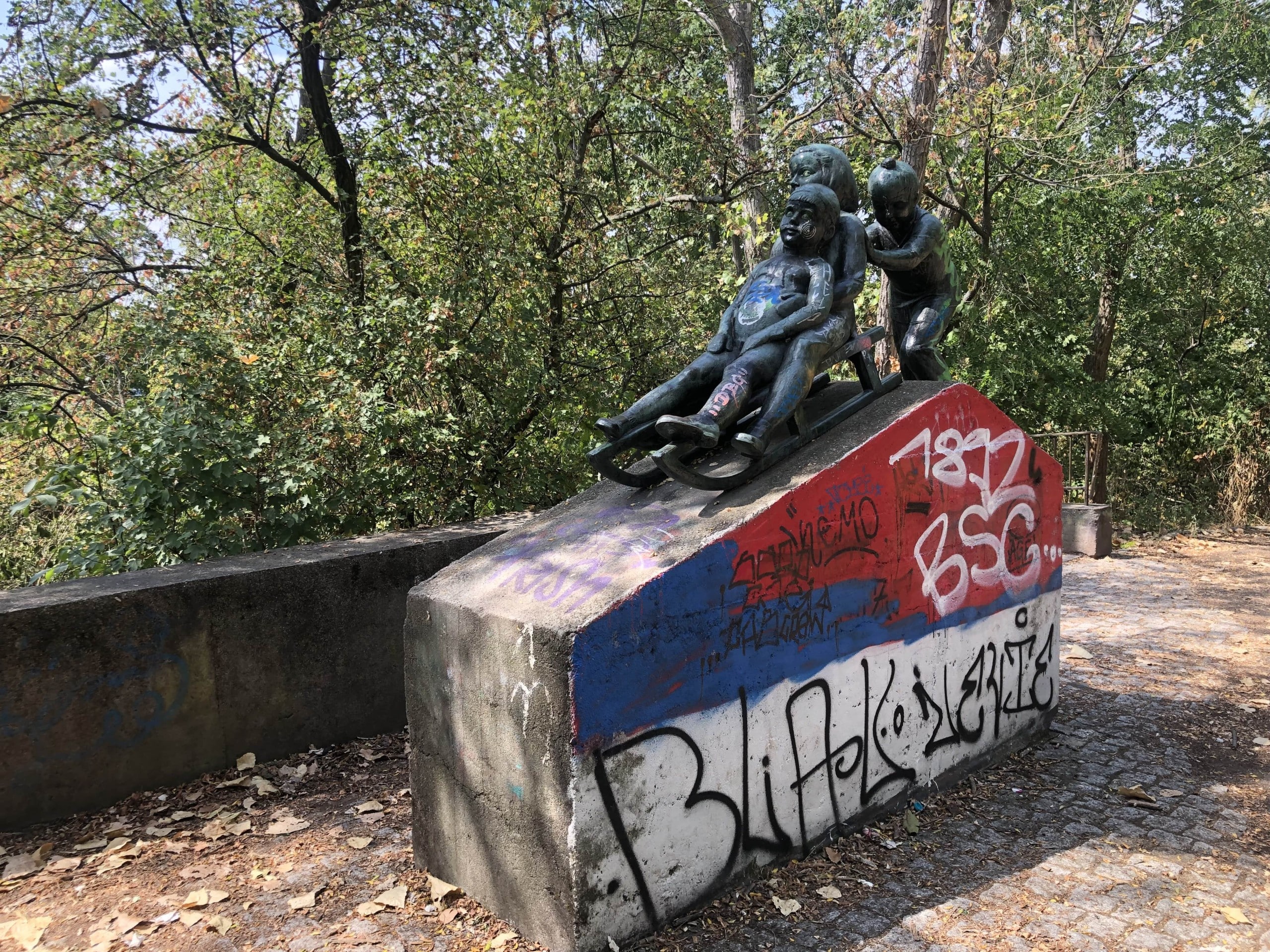 Eine Bronzeskulptur spielender Kinder auf einem Betonstockel, der an eine Rutsche erinnert und der mit Graffitis bemalt ist; im Hintergrund dichter Wald; Volkspark Prenzlauer Berg, Foto: Stefan Reimann