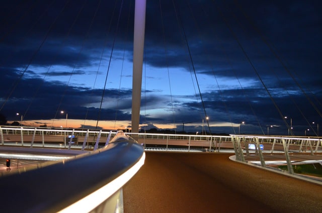 Nachtaufnahme von einer schwebenden runden Brücke. Sie hängt an Stahlseilen. Der Boden ist roter Asphalt.
