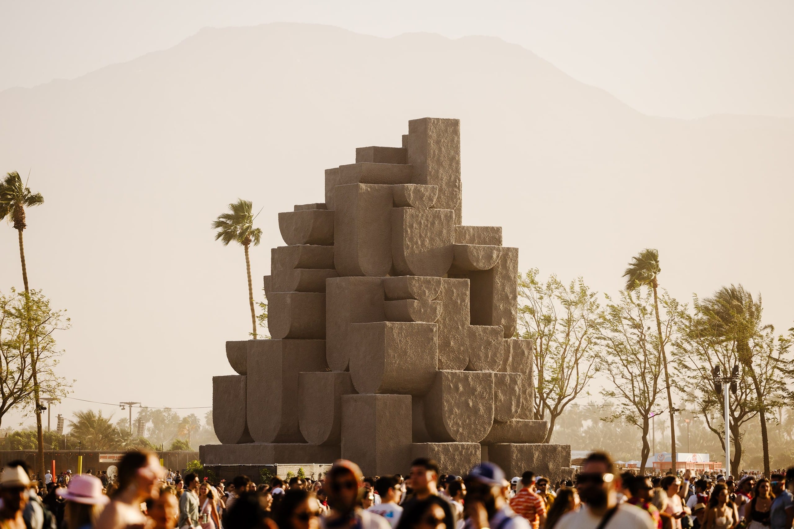 Ein Turm aus halbrunden Blöcken, davor eine Menschenmenge, im Hintergrund Palmen und im Dunst eine Bergkette. Coachella 2024, Nebbia, Babylon, Foto: Lance Gerber