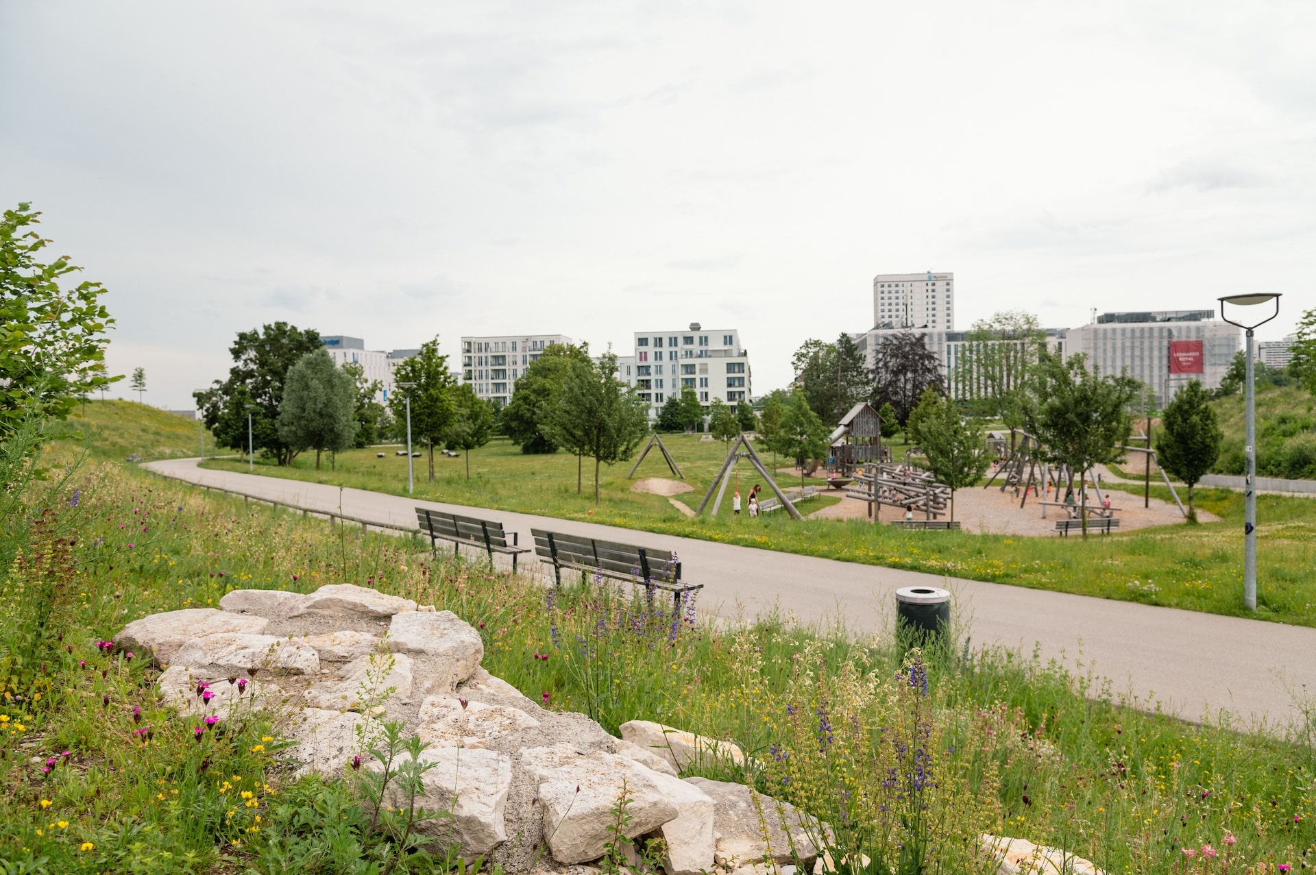 Ein städtischer Park, im Vordergrund Steine in einer hoch bewachsenen Wiese, dahinter zwei Bänke an einem Weg, ein Spielplatz sowie Hochhäuser im Hintergrund. Der Bayerische Landschaftsarchitektur-Preis 2024 geht an den Oberwiesenfeldpark im Münchner Norden vom Büro mahl gebhard konzepte. Foto: © Laura Loewel